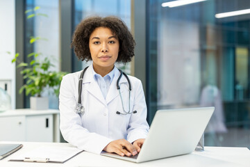 Portrait of a serious young African American female doctor in uniform sitting at a desk, working on a laptop and looking confidently at the camera