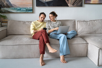Obraz premium Carefree positive married partners husband and wife relaxing on sofa using smartphone and laptop browsing online, scrolling social media and sharing funny videos. Happy couple enjoying time together.