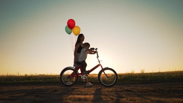 Happy family in park at sunset. kid learns to ride bike in park on green grass. Happy kid on his birthday. Mom helps teach her son to ride bike at sunset in park. Mom helps child. Kid on bike in park