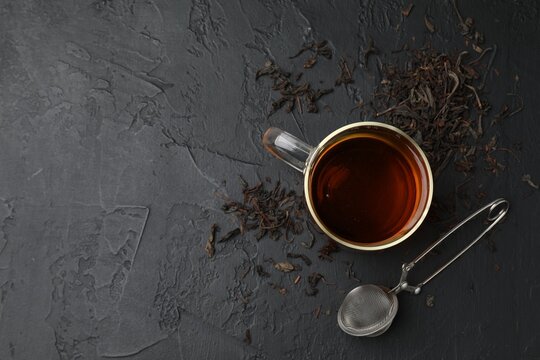 Cup of tea, dry leaves and metal infuser on black table, top view. Space for text