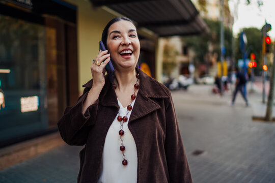 Joyful Hispanic woman laughing on smartphone during street call, reflecting human connection and lighthearted digital communication in vibrant modern freelance lifestyle.