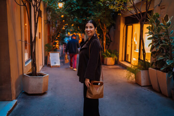 Fototapeta premium Smiling Hispanic woman standing on cozy pedestrian street with warm shop lights, holding shoulder bag, representing evening stroll, relaxation, and friendly urban ambiance.