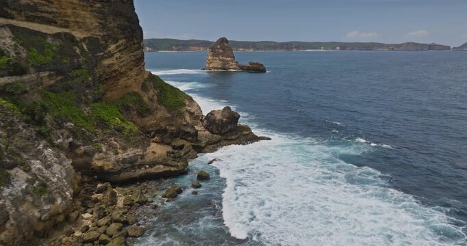 Bukit Merese coastline on Lombok, Indonesia with rugged cliffs, green vegetation, vivid blue sea and waves crashing around iconic Batu Payung offshore rock formation, aerial view drone flight panorama