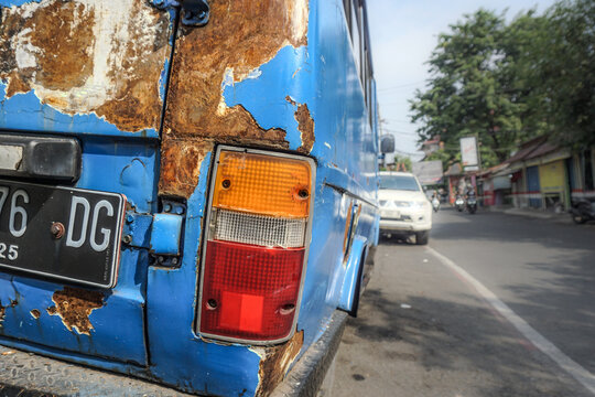 This is a close-up, ground-level photograph capturing the weathered rear of an old, blue bus parked or driving slowly on the side of a street under a bright, slightly overcast sky. The image uses a sh