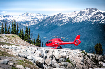 A vibrant red helicopter rests on a rocky outcrop against a backdrop of snow-capped mountains. Conifer trees and blue skies enhance the scenic view. © Ron