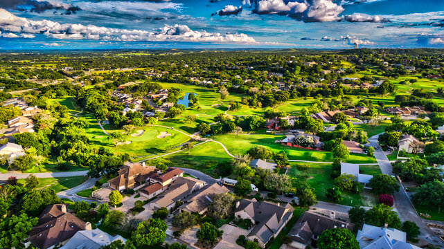 An expansive aerial view showcases a residential community featuring luxury homes nestled amongst lush green trees under a vibrant blue sky, perfect for real estate or urban development features.