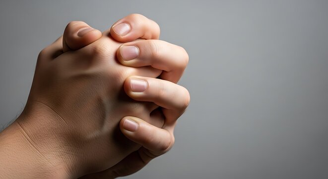 Close-up of two human hands clasped together, possibly in prayer or deep contemplation, against a soft, blurred grey background with subtle lighting.