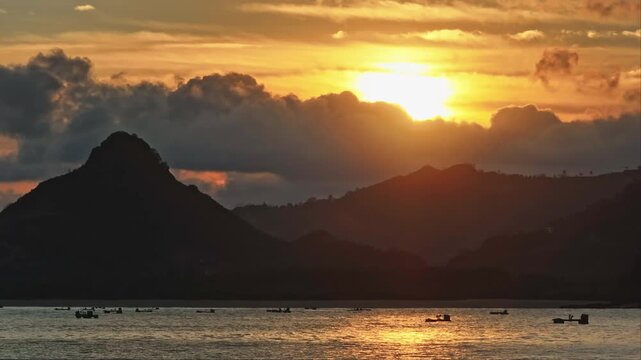 Selong Belanak Beach sunset illuminates the ocean as fishing boats drift along the water, with silhouetted Lombok mountains and golden clouds framing the horizon. Nature landscape. Natural background