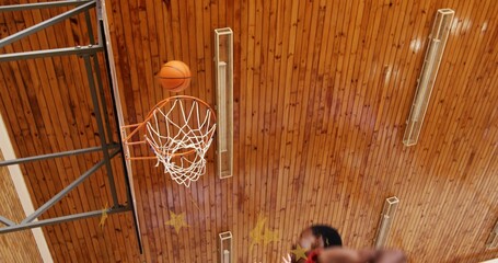 Naklejka premium Sinking orange basketball falling through rim in school gymnasium with players' jerseys and hands