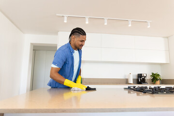 African American man cleaning kitchen island countertop in modern kitchen with yellow gloves, cloth