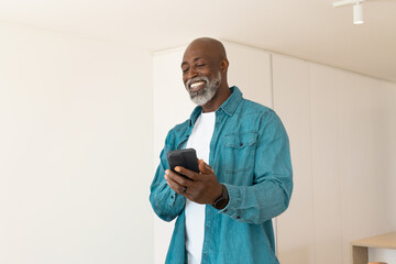 Senior African American man wearing denim shirt and smartwatch holding smartphone smiling at home © wavebreak3