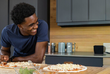 African American man leaning on wooden counter, preparing 2 pizzas with tomato sauce, basil, cheese
