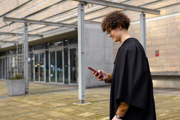 Man in academic gown walking across plaza under glass canopy, checking red smartphone, copy space © wavebreak3