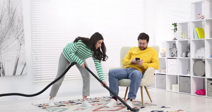 Gender inequality concept. Man with smartphone resting in armchair while his wife doing household chores indoors