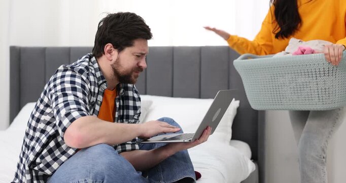 Gender inequality concept. Man using laptop on bed while his wife doing household chores indoors