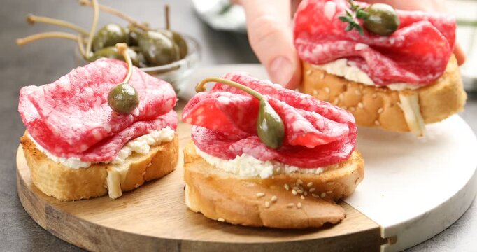 Woman taking delicious bruschetta with salami sausage, cream cheese, thyme and capers from board at grey table, closeup