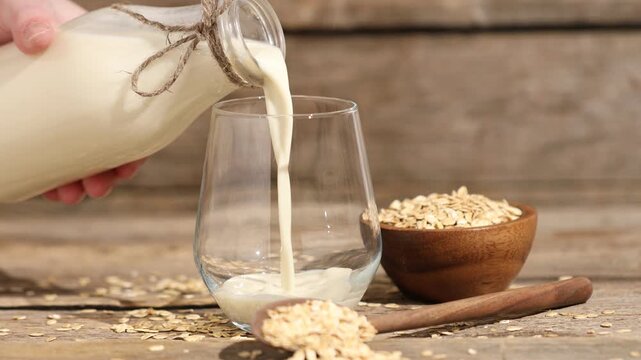 Woman pouring oat milk into glass and flakes at wooden table, closeup