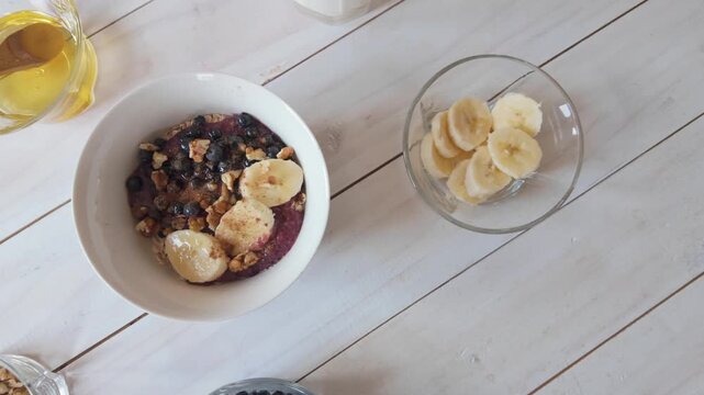 Slow motion left to right camera movement over oatmeal bowl with banana, blueberries, walnuts and honey on bright wooden surface