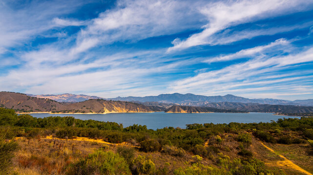 A wide panoramic view of Lake Cachuma in California, framed by rolling golden hills and the Santa Ynez Mountains beneath sweeping blue skies and high cloud formations.