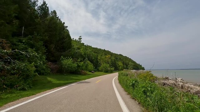 Slow-motion POV cycling a curve on M-185, Mackinac Island, MI. The paved path follows the rocky Lake Huron shore with green forest and cyclists ahead under a clear summer sky.
