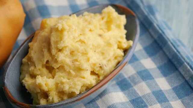 Close-up cinematic video of creamy mashed potatoes in a bowl on a checkered blue and white tablecloth, with a fresh potato nearby, showcasing homemade comfort food