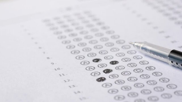 Close up of black ballpoint pen resting on filled bubble test sheet with blurred background showing multiple choice questions