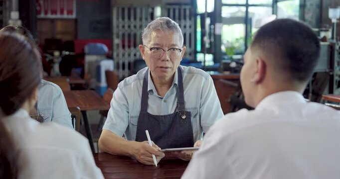 Asian senior cafe owner shares ideas with young employees, teamwork collaboration between generations creates trust, improves workflow, and supports family small business success in friendly workspace
