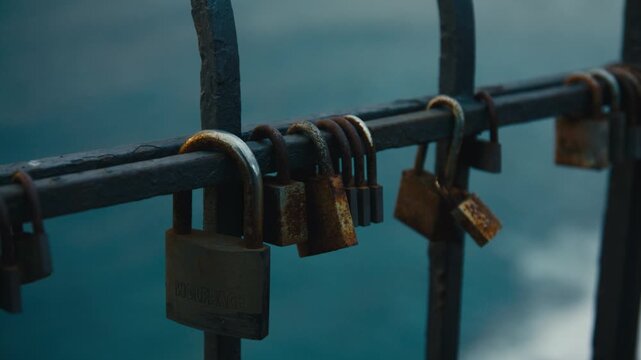 Close-up of weathered and rusted love padlocks attached to an iron seaside fence, shallow depth of field with soft blue ocean backdrop at dusk.