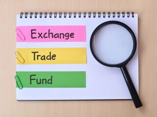 Exchange Traded Fund (ETF) handwritten on notebook with magnifying glass with wooden background. Concept of investing, financial planning, and stock market analysis