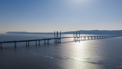 Obraz premium Cable stayed bridge with tall towers and a lengthy viaduct section extending across a wide river, reflecting the bright sky and forming long shadows on the water