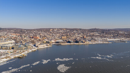 Obraz premium Aerial view capturing a vibrant urban waterfront with modern buildings and snow dusted hills, while ice floes drift across the cold river under a clear winter sky