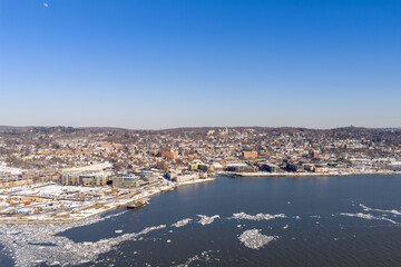 Obraz premium Winter aerial view showing urban development along a frozen riverfront, featuring many buildings, snow dusted hills, and scattered ice chunks floating on the water under a clear blue sky