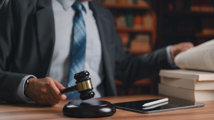 Professional male lawyer wearing suit holding wooden gavel reading heavy legal books sitting inside library office desk near modern digital tablet pen reviewing important court justice documents very