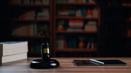 Wooden desk displays black judge gavel beside modern digital tablet stylus pen stacked legal books inside dark library room featuring blurred bookshelves behind representing modern justice system © Mdisk