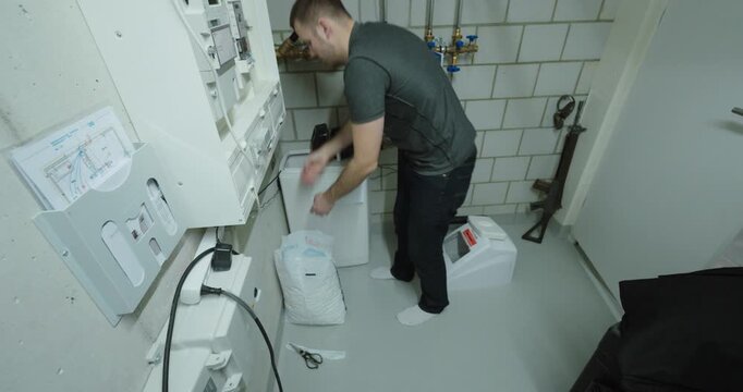 Man pouring salt tablets into a home water softener unit in a technical room, demonstrating routine plumbing maintenance, limescale prevention and residential water treatment care.