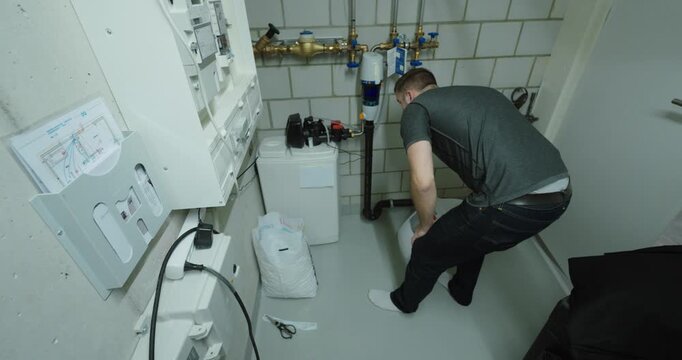 Wide-angle view of a technician inspecting and refilling a domestic water softener unit in a utility room, highlighting plumbing maintenance, water treatment systems and household infrastructure care.