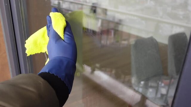 Person wearing blue rubber gloves wiping glass window with yellow microfiber cloth for cleaning service