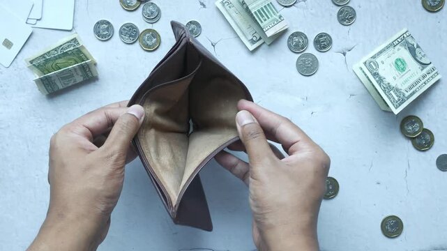 Top view of hands opening empty brown leather wallet surrounded by cash and coins
