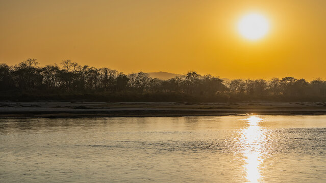 Sunset over the river in the jungle. The sun is low. The sky is orange. Trees on the shore. A sunny path on the water. Nepal. Rapti River. Chitwan National Park.
