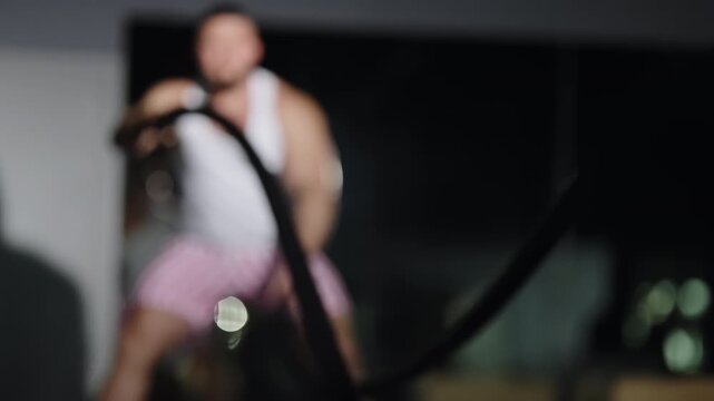 Out-of-focus video frames show an athlete driving intense battle rope training in a dim indoor gym, highlighting powerful motion, strength conditioning, and rapid arm waves through a low-light workout