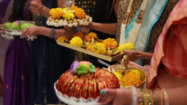 Women holding traditional offering plates with marigold flowers and ceremonial items during Indian wedding ritual. Close-up of cultural preparation in colorful ethnic attire.