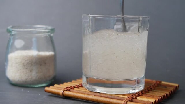 Close up slow motion shot of stirring psyllium husk in a glass of water, next to a jar of psyllium husk, with gray background, healthy food concept