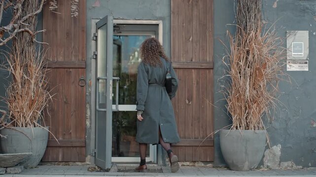 Caucasian florist approaching shop entrance, hands on handle pushing glass door interior glimpse warm lights preparations arranging stems morning routine small business atmospheric shot boots coat