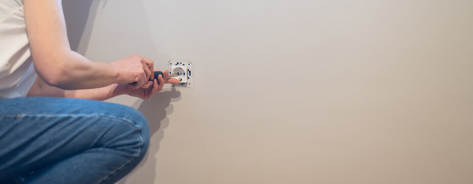 Man crouching installing a new electrical outlet into a light-colored wall using a screwdriver during home renovation or diy electrical repair and wiring work