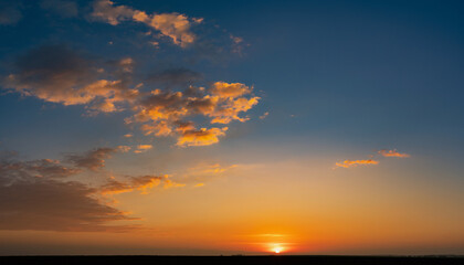 Obraz premium Beautiful sunset with sun on horizon, bright orange glow illuminating scattered clouds against gradient blue and orange sky, dark landscape silhouette