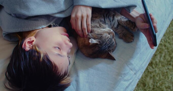 A teenage girl lies comfortably in bed, intently scrolling through her smartphone while her cat relaxes peacefully beside her, sharing a quiet and cozy moment together.