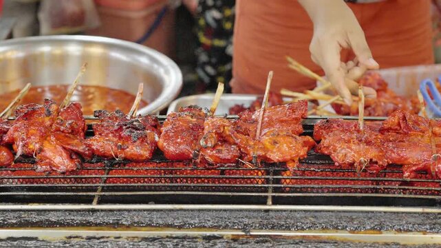 Close-up footage of chicken skewers grilling over a hot charcoal stove at a Thai street market. A vendor flips the skewers by hand, showcasing authentic street food cooking and smoky outdoor atmospher