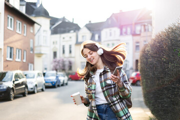 Fototapeta premium Happy woman in headphones with smartphone dancing on town street. Girl listening to music playlist outdoors, walking in city, having fun. Urban student lifestyle. Whimsical, zest for life