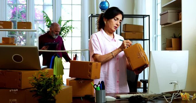 Indian senior couple doing E-commerce business from home, busy working in office managing online orders, packing parcels, dispatching boxes, taking customer calls and handling inventory together