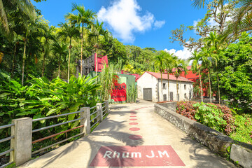 Fototapeta premium Martinique, French Antilles - Dec 27, 2025: Rhum JM distillery buildings nestled in lush tropical vegetation with palm trees and a small stream reflecting a bright blue sky